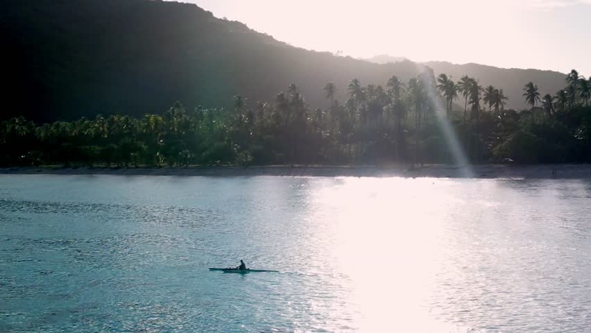 Lone person paddling a kayak across a calm turquoise sea toward a palm fringed tropical island