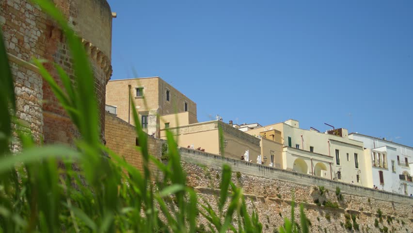 Historic defensive walls rise above coastal vegetation in Brindisi, Apulia, Italy, highlighting centuries of military architecture beneath a clear Mediterranean sky, static low angle shot.