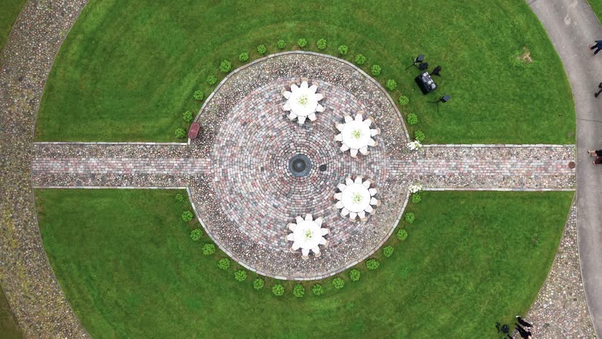 Aerial footage of a symmetrical circular brick pathway with four large white floral sculptures, surrounded by lush green lawn in daylight. Overhead shot offers clear, geometric composition.