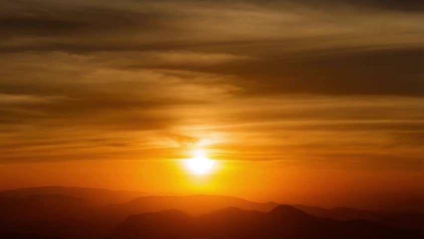 Beautiful cinematic landscape view of a brilliant orange sun setting on the horizon over silhouette mountain ranges. Soft wispy clouds catch the warm golden hour light, perfect for wilderness travel, inspiration, nature, and peaceful dawn concepts.