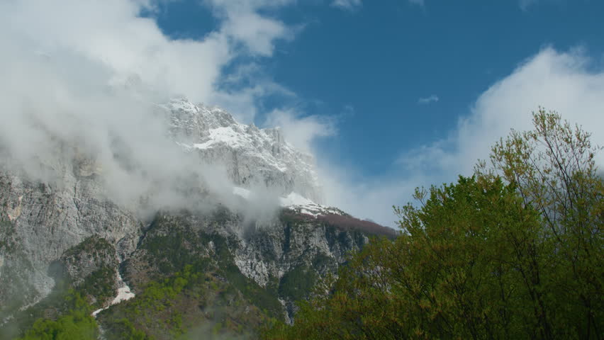 Snowy mountain peak valley forest and scattered clouds, static tripod view. Alpine summit with snow, rocky cliffs, green slopes and trees foreground.