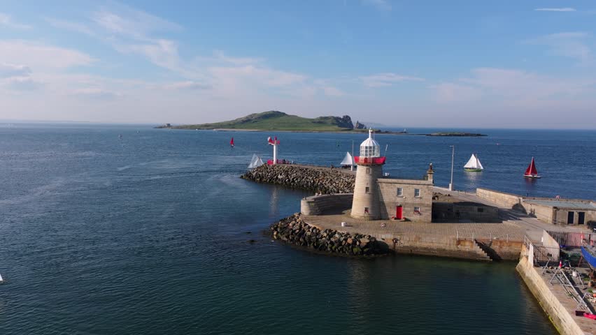 Sailing dinghies glide past a red and white lighthouse jetty with Ireland’s Eye island in the background under clear skies.