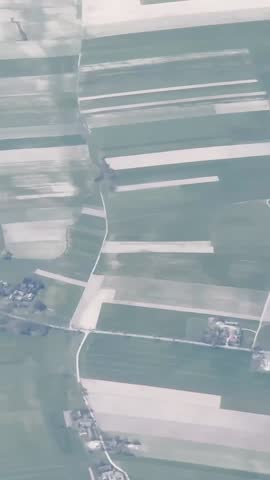 Vertical aerial view of patchwork farmland and a small village through haze, with pale crop strips, country roads and geometric field patterns.