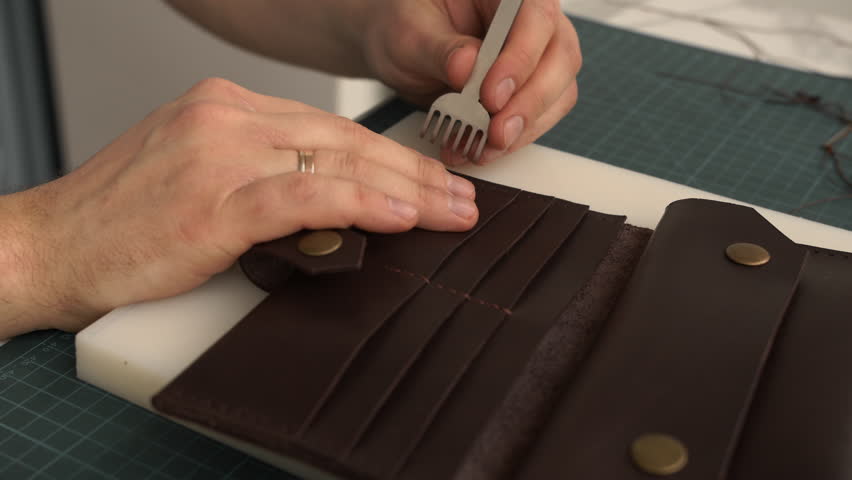 Craftsman punching stitching holes in a handmade brown leather wallet using a pricking iron tool