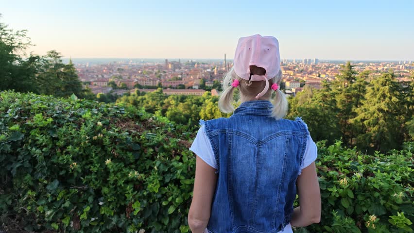 Woman standing on a hill viewing the panoramic cityscape of Bologna, Italy, with historic buildings and the famous Two Towers, Asinelli and Garisenda, under a clear sky during a summer evening