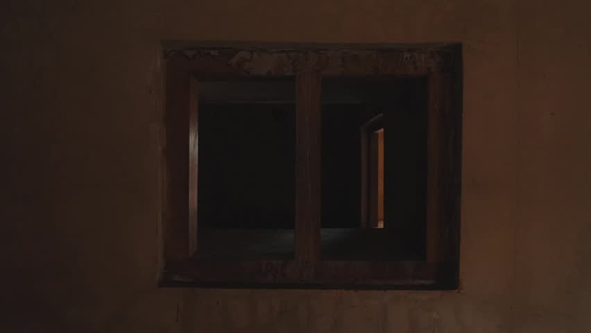 A close-up shot of an old weathered wooden window frame in a dark room. Warm light illuminates the interior edge, creating a moody, dramatic, and suspenseful atmosphere.