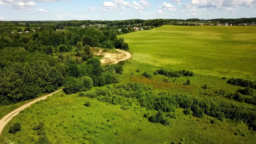 Aerial view of a rural landscape with a winding dirt road, lush green fields, and a village in the distance. Car on the dirt road adds a focal point for transport and accessibility.