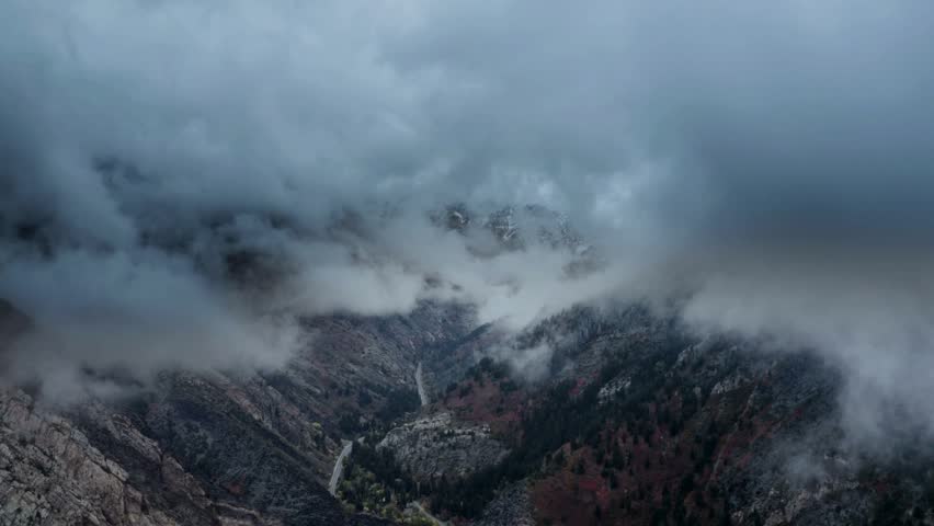Winding road snakes through a rugged valley beneath moody blue clouds. Snow-dusted peaks emerge through dense fog above rocky slopes.