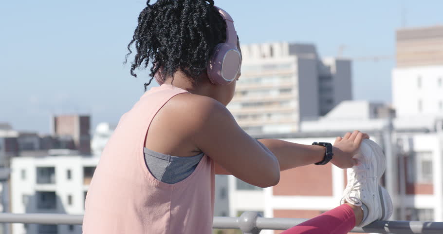 Woman placing right leg on rooftop railing, reaching and gripping shoe, warming up for run. Stretching, fitness, headphones, urban, skyline, athlete, balance