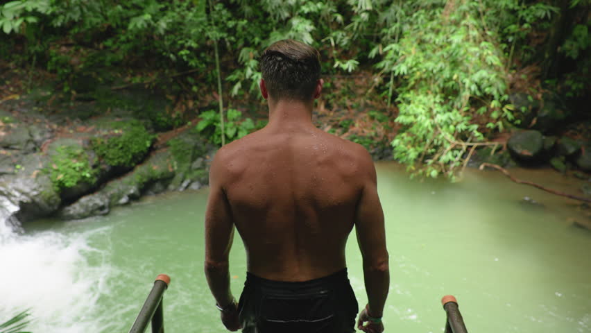 Slow motion footage captures athletic young man performing backflip dive from platform into natural green pool below tropical waterfall surrounded by lush jungle vegetation and rocky cliffs.