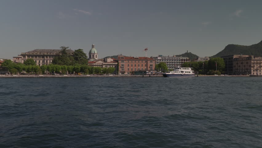 View from the lake of the city of Como, in northern Italy. An elegant natural amphitheater nestled between the crystal-clear waters and the surrounding mountains. The pastel-colored facades of the historic buildings and the cathedral and bell towers emerges harmoniously among the buildings of the center. Along the lakefront, centuries-old trees, manicured gardens, and elegant villas create an atmosphere of great charm.