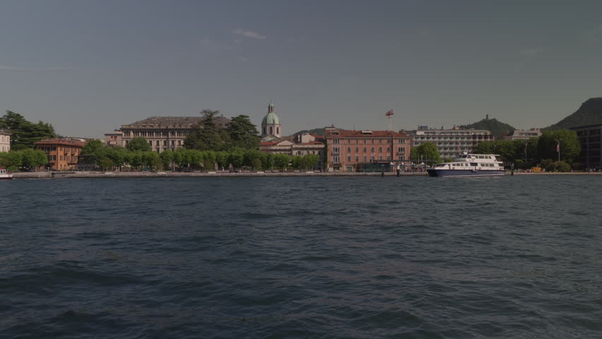 View from the lake of the city of Como, in northern Italy. An elegant natural amphitheater nestled between the crystal-clear waters and the surrounding mountains. The pastel-colored facades of the historic buildings and the cathedral and bell towers emerges harmoniously among the buildings of the center. Along the lakefront, centuries-old trees, manicured gardens, and elegant villas create an atmosphere of great charm.