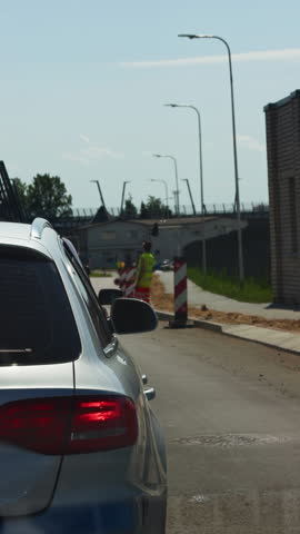 Silver car waiting beside roadworks with traffic worker, barrier posts, and urban street under bright daytime sky