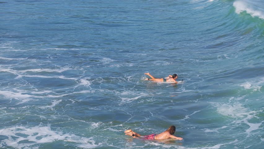 Surfing scene shows two people paddling on surfboards through turquoise ocean waves. White foam spray from breaking swells surrounds them as they navigate the blue water on a bright sunny day.