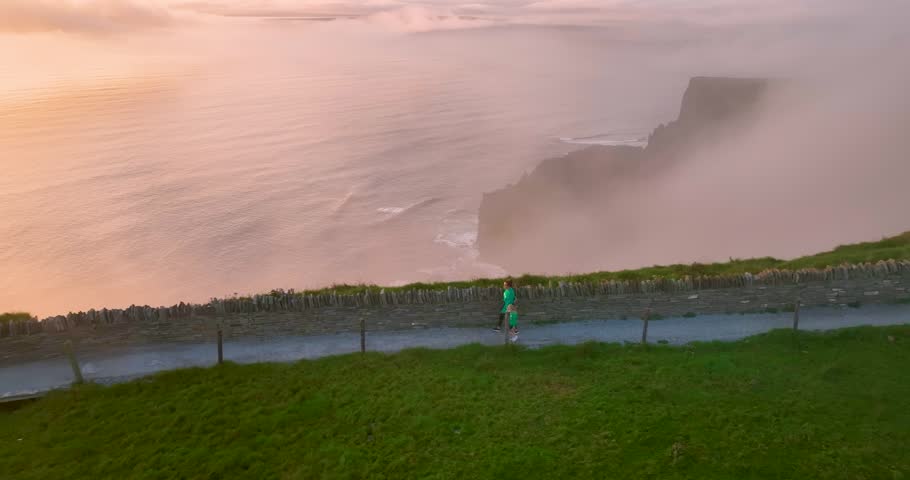 Tourists walking along Cliffs of Moher coastal trail during misty sunset in County Clare, Ireland