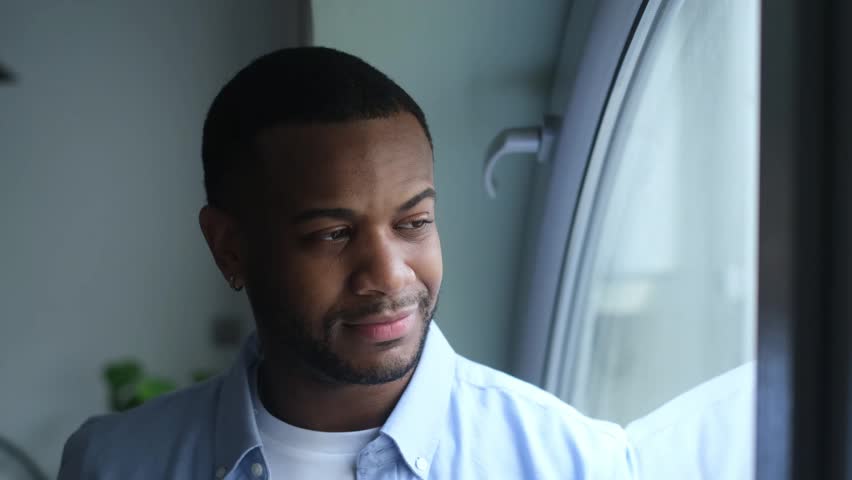 A happy and thoughtful young African American man gazes out a window with a warm smile. His positive expression conveys contentment and reflection in an indoor setting.