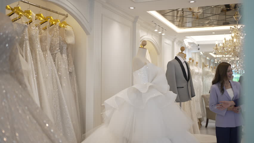 Tailor touches the hem of a glittering wedding dress while wearing a tape measure, discussing details with a customer nearby.