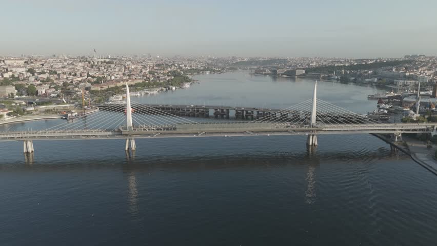 Istanbul, Turkey. The Golden Horn Metro Bridge spans the Halic estuary in warm morning sunlight, its twin white pylons and harp-arranged cables forming a striking cable-stayed structure. The bridge carries the M2 metro line and features the unusual Halic Metro Station integrated directly into its central span, a design that places platforms above the water between the Beyoglu and Fatih districts. An aerial drone perspective reveals the bridge