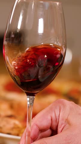 A person swirls red wine in a glass over a table with pizza and appetizers.