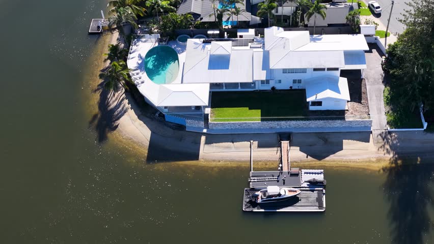 High-angle drone shot orbits a modern white waterfront estate featuring a pool and private dock.