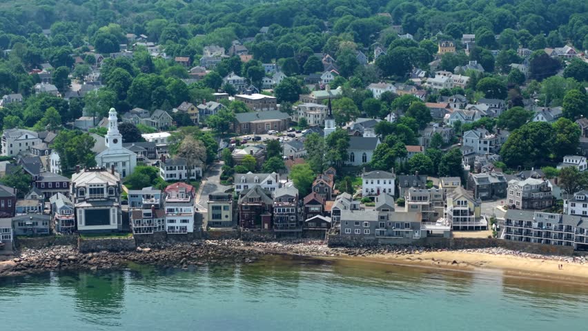 Quaint harbor town spreads along rocky New England coastline, where dozens of moored boats fill protected bay surrounded by historic homes in Rockport. Massachusetts, USA. Aerial drone wide shot