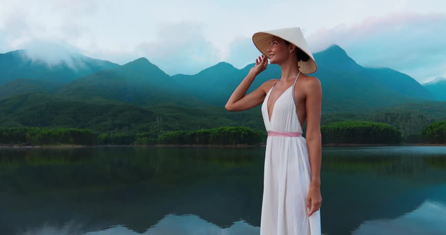 Static medium shot of a young woman in a white dress and straw hat posing at Tra Ly Lake, Thai Binh, Vietnam. Misty mountains reflect on calm water. Ideal for travel and tourism campaigns.