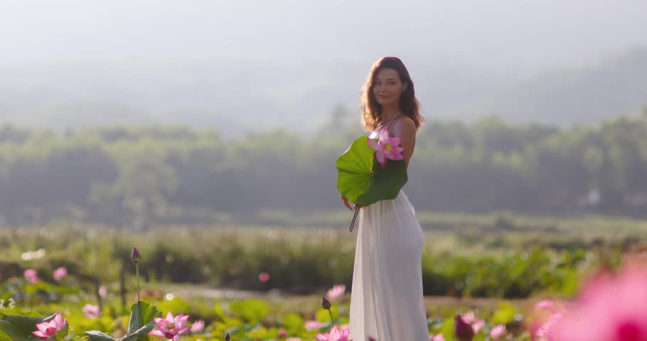 Young woman in flowing white dress smells and holds pink lotus blooms in misty morning pond, Tra Ly, Quang Nam, Vietnam. Dreamy, peaceful mood. Perfect for wellness, beauty, and lifestyle campaigns.