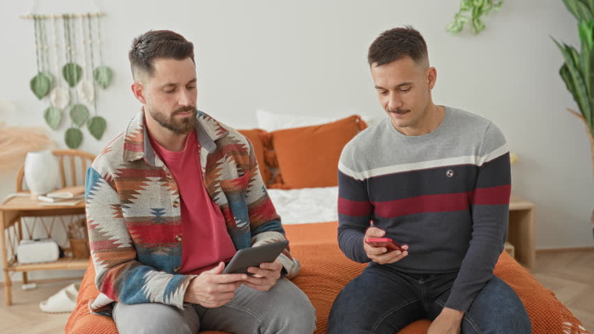 Two men sit on a bed holding a tablet and smartphone, pointing at the screen in a bright bedroom; affection comfort connection.