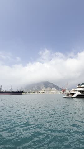 ANTALYA, TURKEY - JULY 2026: Vertical wide shot of a commercial harbor and shipyard with cargo ships, yachts, and industrial cranes against mountains partially covered by clouds.