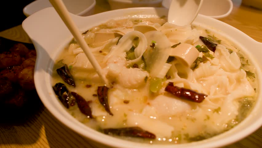 Close up shot of chopsticks picking up fish and noodles from a spicy ceramic bowl.