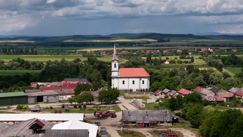 A traditional Hungarian village church stands prominently amidst a green rural landscape under a dramatic cloudy sky, portraying serene country life.