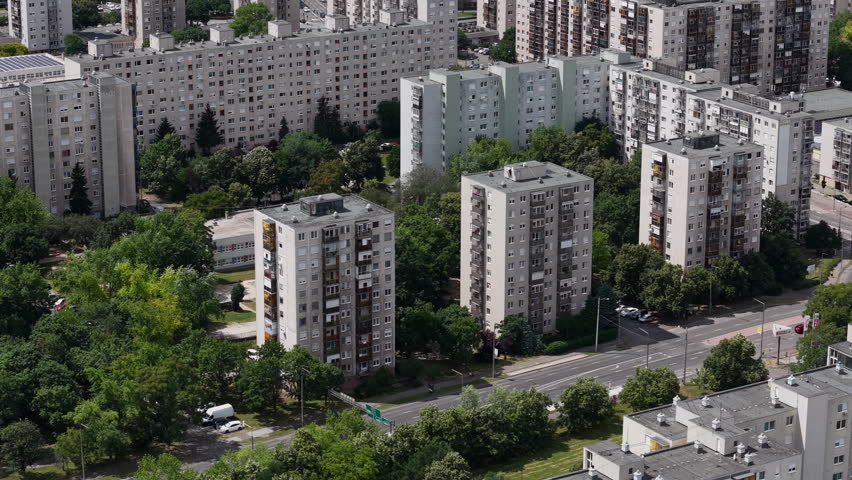 An aerial perspective shows high-rise residential apartment blocks surrounded by lush green trees and urban infrastructure in Budapest, Hungary.