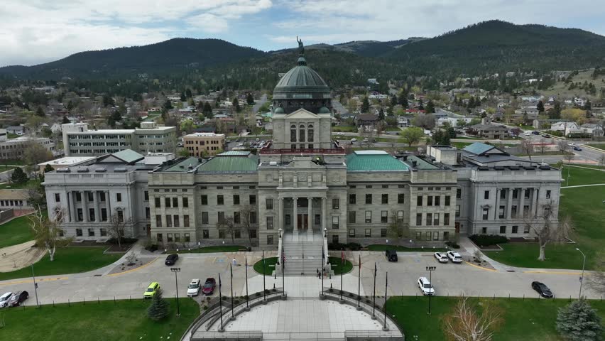 Montana State Capitol dome rises above Helena skyline, framed by government buildings, tree-lined streets and forested mountains under cloudy sky. Helena, Montana, USA. Aerial drone backwards shot