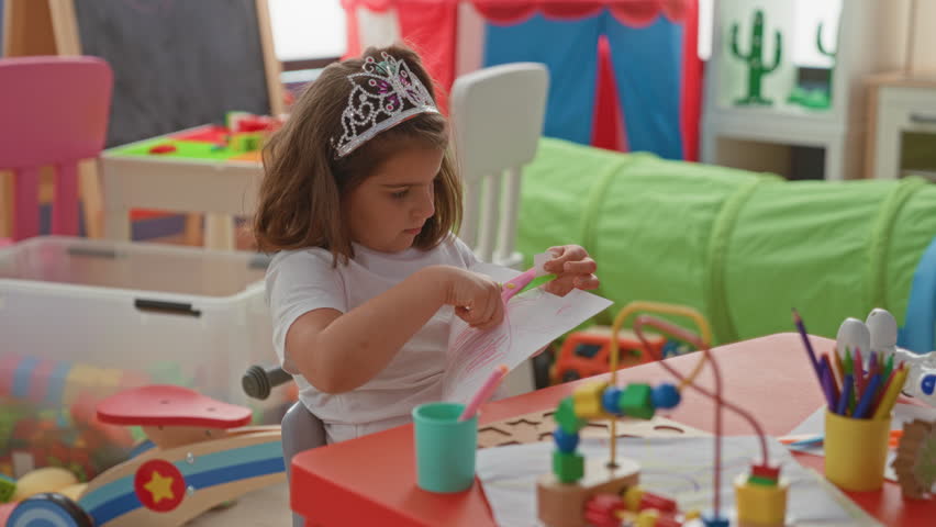 Girl cuts paper with scissors at a play table inside a building wearing a tiara and using hands to snip shapes; concentration creative play.