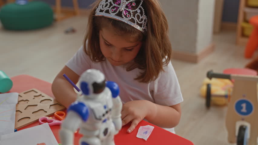 Little girl wearing a jeweled tiara draws on paper with a purple pencil, right hand on a red table beside a toy robot in a playroom building; creative concentration.