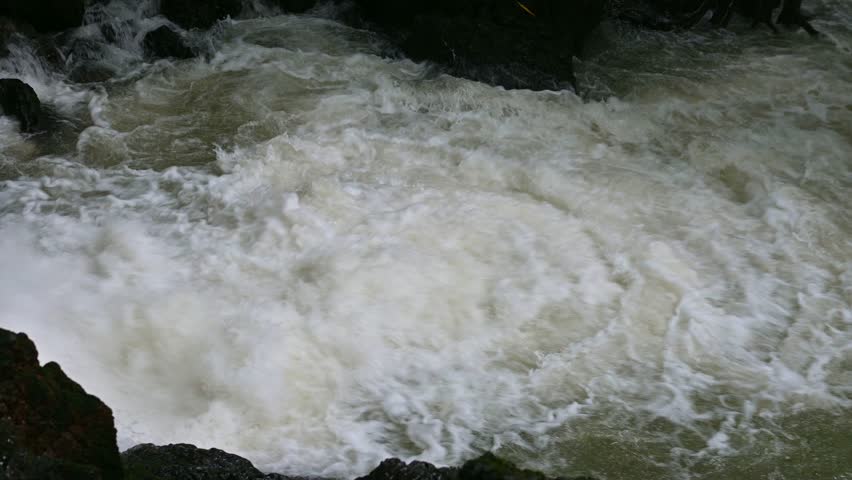 Close up of violent rapid river stream with white foam splash. Water surface background. Powerful rushing water through rocks in a mountain. 