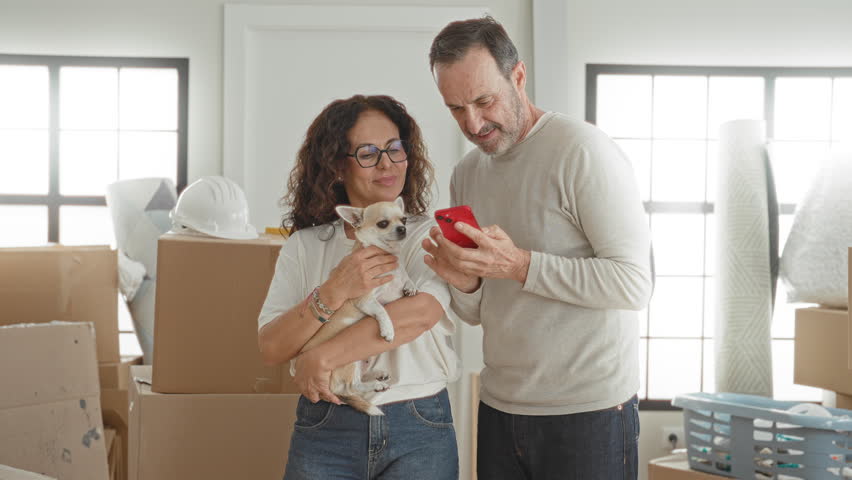 Man fist pump and woman holding small dog as they laugh viewing a red smartphone amid packed boxes in a building; moving day joy.