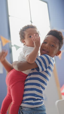 Older brother holds baby sister in his arms by a tall window and colorful bunting in a playroom building; affection.