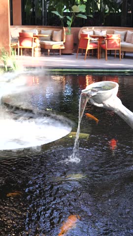 Carved stone spout pouring steady water into reflective resort pond with rising mist, gentle ripples, sunlit lounge seating, tropical foliage, and tranquil modern hospitality setting