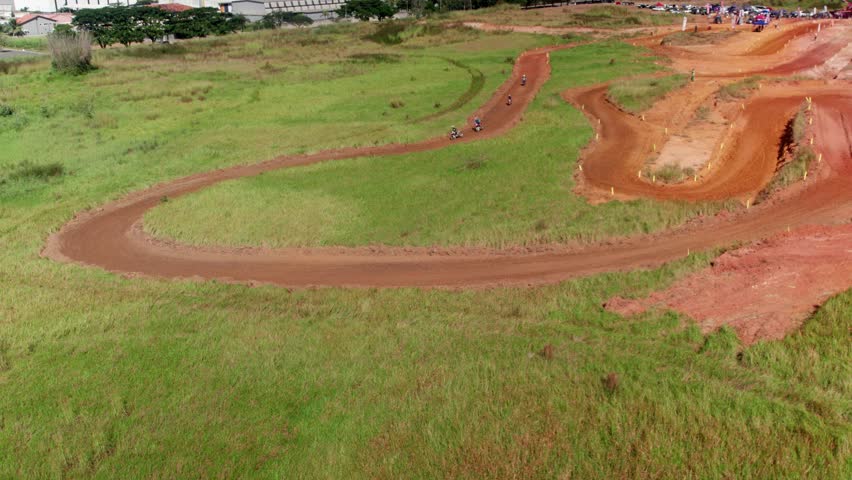 Aerial drone shot showing several motocross riders racing and navigating a long curve on a dirt track. The sunny outdoor scene features green grass and an event area in the background.
