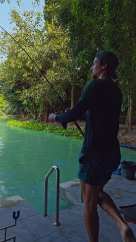 Vertical video. Man Holding Bent Fishing Rod While Fighting Strong Fish Near Lake Shore Side View Outdoor Action Angling Sport Tension Moment.