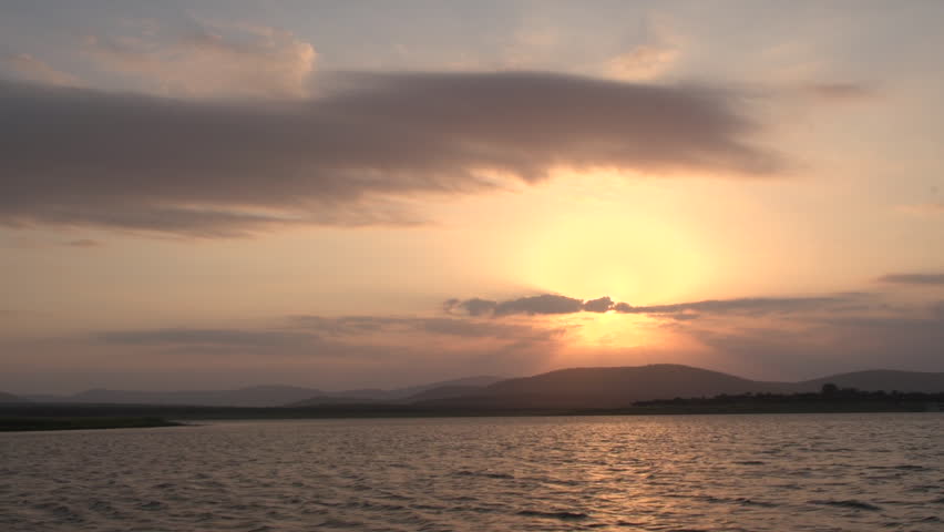 Spectacular wide shot of a glowing golden sunset over a large rippling lake, with sun rays bursting through clouds behind a beautiful silhouette of rolling mountain ridges in South Africa.