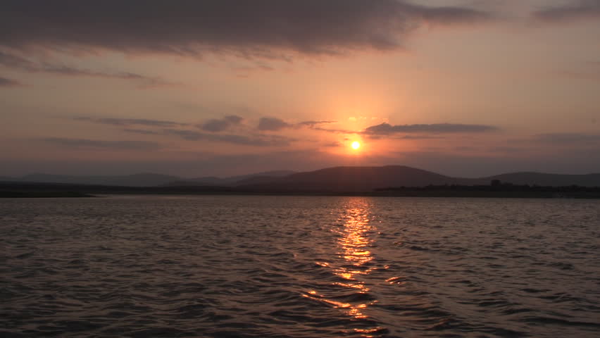 Spectacular wide shot of a glowing golden sunset over a large rippling lake, with sun rays bursting through clouds behind a beautiful silhouette of rolling mountain ridges in South Africa.