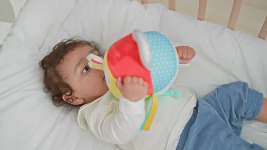 Girl holds colorful plush ball with small hand while lying on white mattress in crib inside building, gazing at the toy; curiosity bonding.