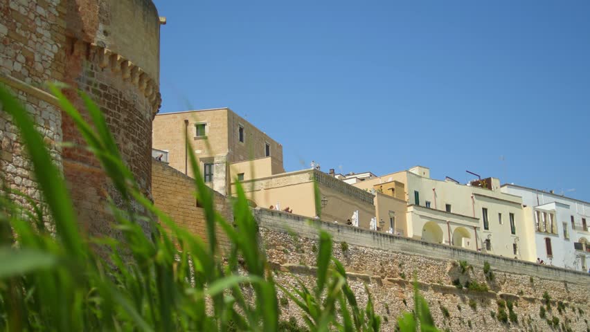 Historic limestone fortifications and traditional buildings overlook coastal vegetation in Brindisi, Apulia, Italy, showcasing centuries of Adriatic military architecture, static low angle shot, windy