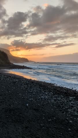Dramatic sunset over exotic volcanic black sand beach with breaking ocean waves and rocky cliffs in Tenerife, Canary Islands, Spain. Beautiful coastal landscape during golden hour twilight.