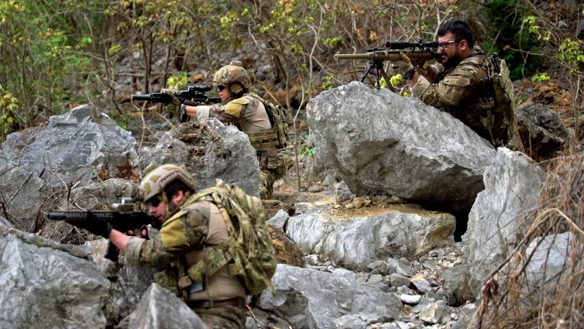 Three armed soldiers in camouflage take cover among large rocks, aiming rifles during a tactical operation in rugged, forested terrain.