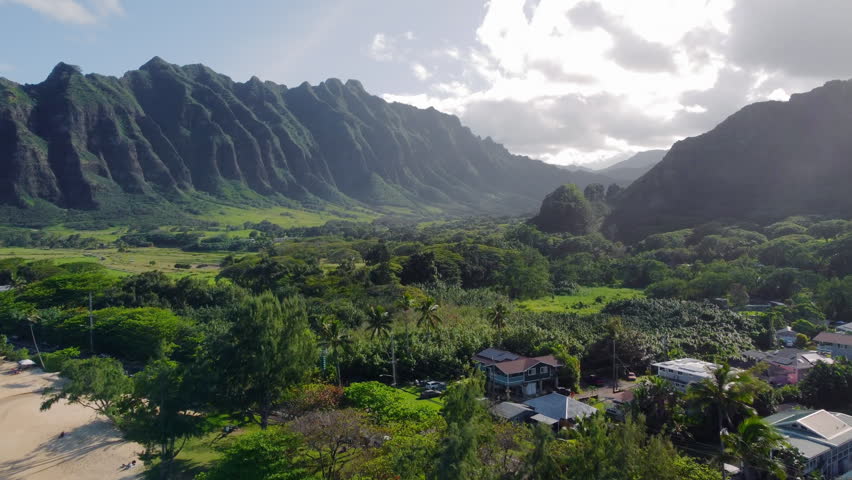 Green Hawaii mountain valley with steep volcanic cliffs and clouds, drone video frame over tropical forest, homes and sunlit island landscape