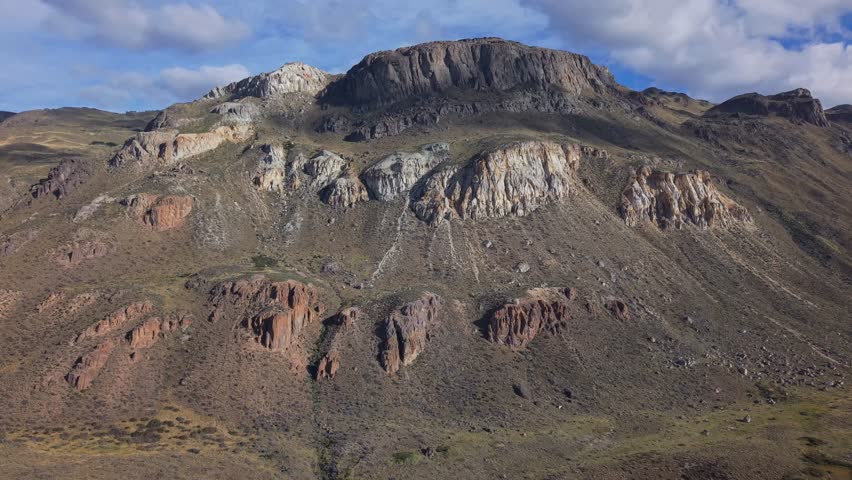 Drone footage gliding past colorful layered rock formations and rugged mountain cliffs under beautiful clouds, showcasing a vast arid landscape with striking geological textures.