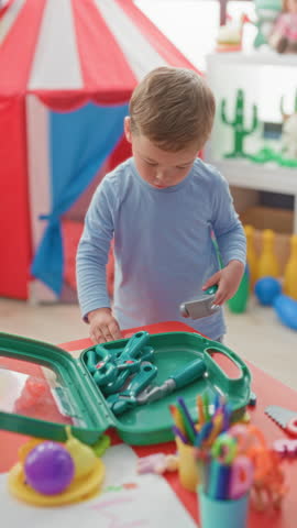 Little blond boy holding a toy hammer and picking tools from a green plastic toolset at a play table in building; concentration curiosity.